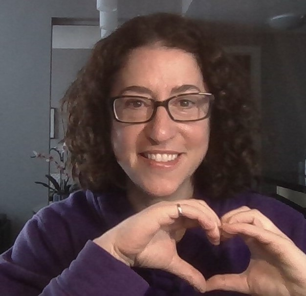Stacie smiling and making a heart shape with her hands, wearing a purple sweater